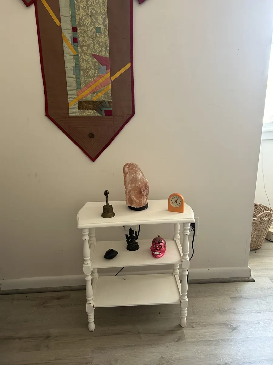 A small white shelf in Kara's office with a salt lamp, a bell and a clock beneath a quilted wall hanging