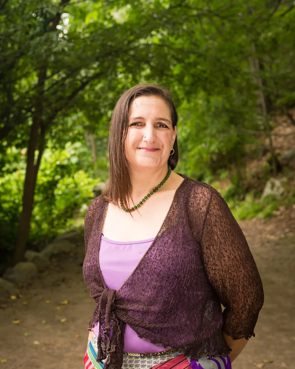 Kara Katz standing in a green wooded clearing, smiling, in a purple shrug and colorful patterned skirt